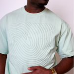A man wearing a pale mint green crewneck t-shirt with a large, raised abstract wave pattern across the chest, paired with a gold watch.
