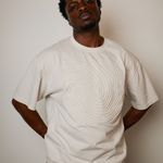 A man wearing a cream-colored short-sleeve t-shirt with a raised, textured spiral pattern on the front, posed against a plain light beige wall.