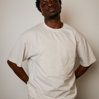 A man wearing a cream-colored short-sleeve t-shirt with a raised, textured spiral pattern on the front, posed against a plain light beige wall.